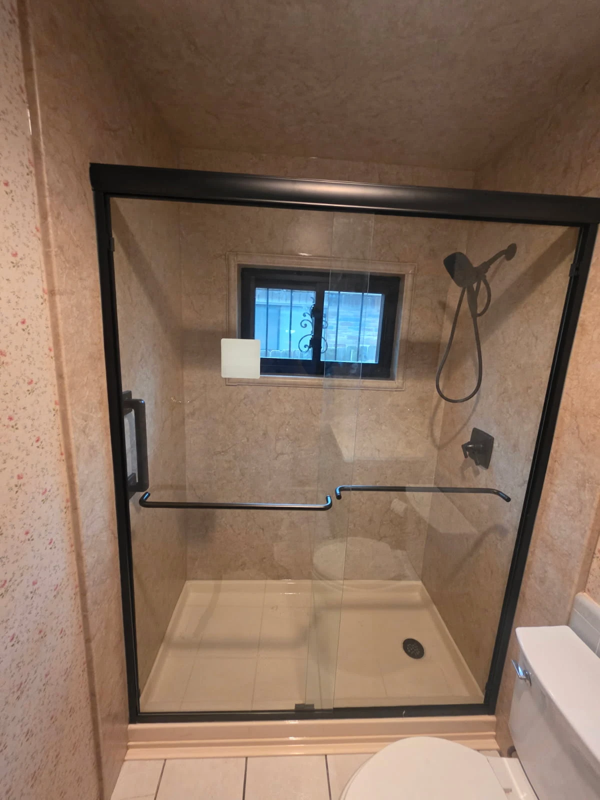 Beige marble-pattern walk-in shower with black-framed bypass glass doors, oil-rubbed bronze handheld and dual grab bars over a low-profile pan