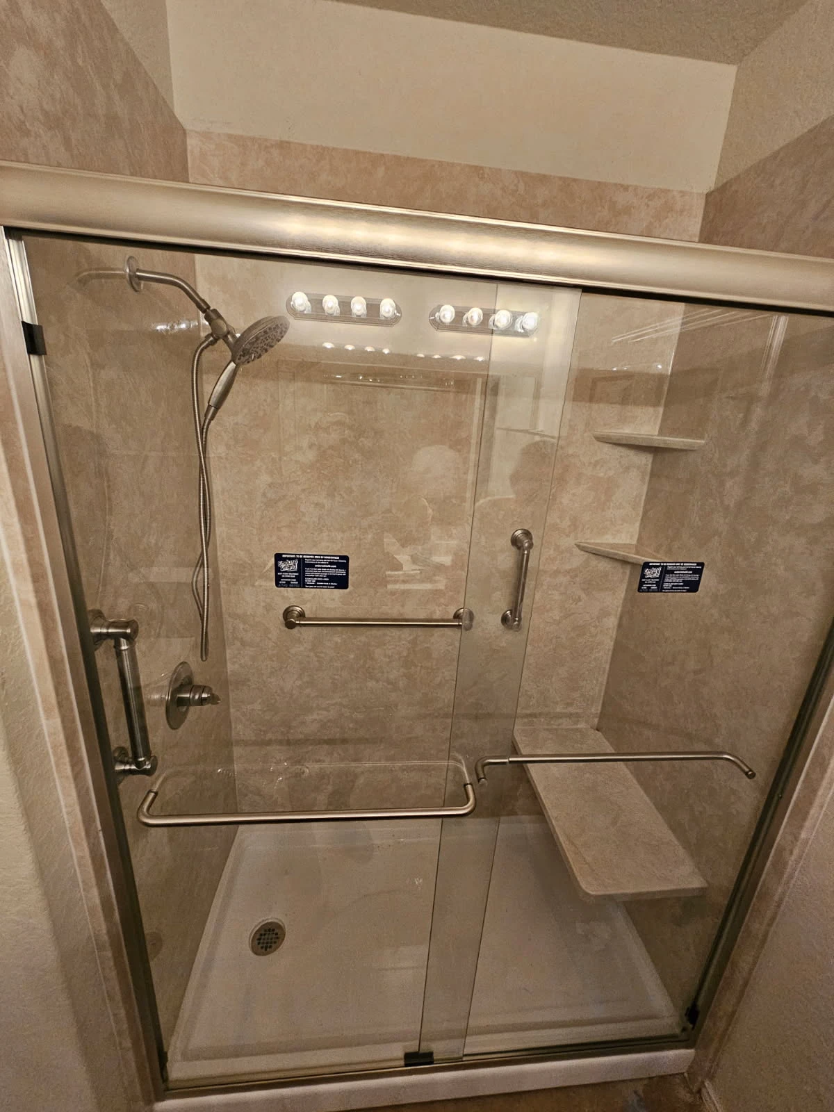 Beige solid-surface walk-in shower with built-in granite bench, multiple grab bars and corner shelves — full aging-in-place setup