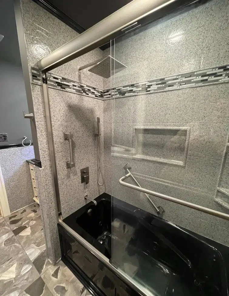 Black soaking tub with granite-pattern surround, glass mosaic accent band, frameless glass shield and brushed-nickel grab bars